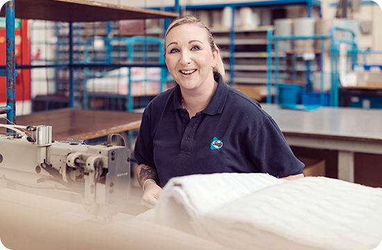 A smiling worker in a textile manufacturing facility standing beside industrial sewing equipment and folded fabric