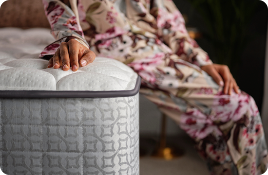 Person sitting beside a plush mattress with hand resting on the mattress edge